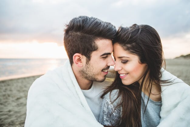 loving-couple-sitting-on-a-beach-covered-by-a-white-blanket_23-2147595916